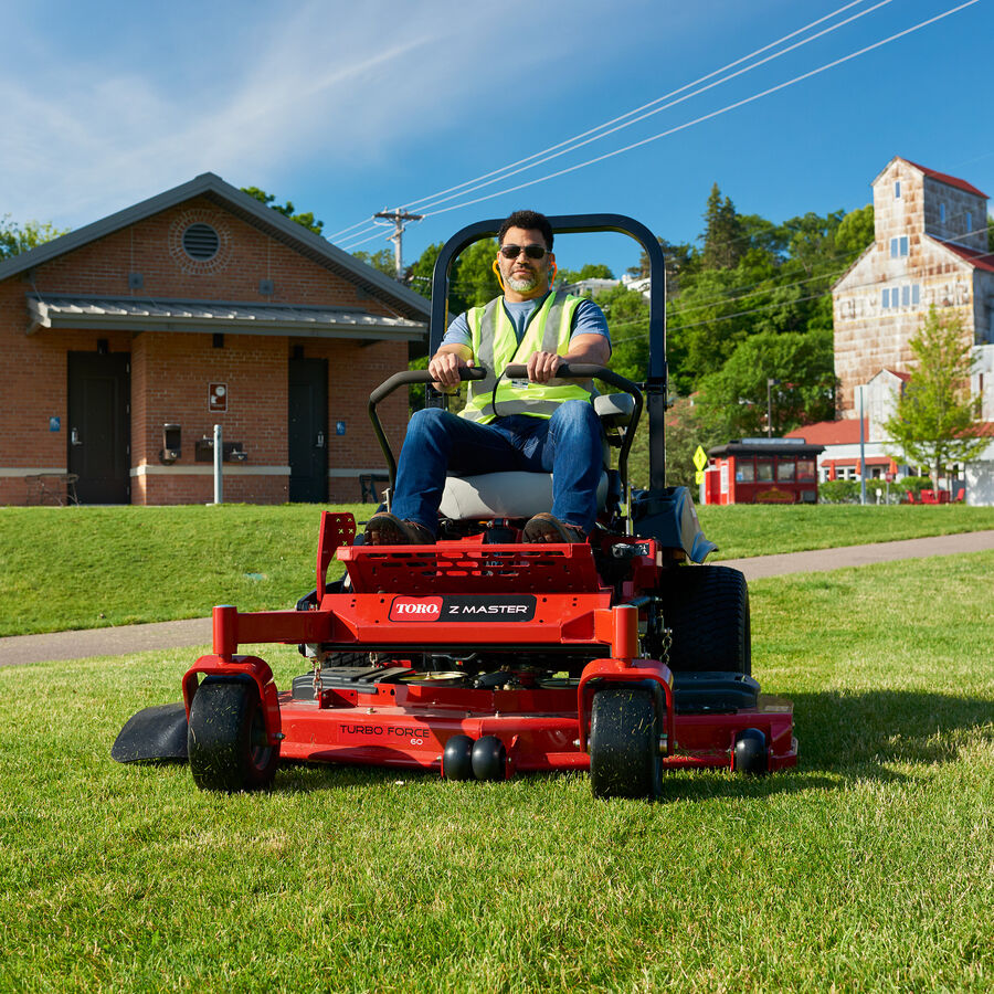 Man on a Toro Z Master 6000 series mower mowing the lawn of a park. There is a few buildings in the background.