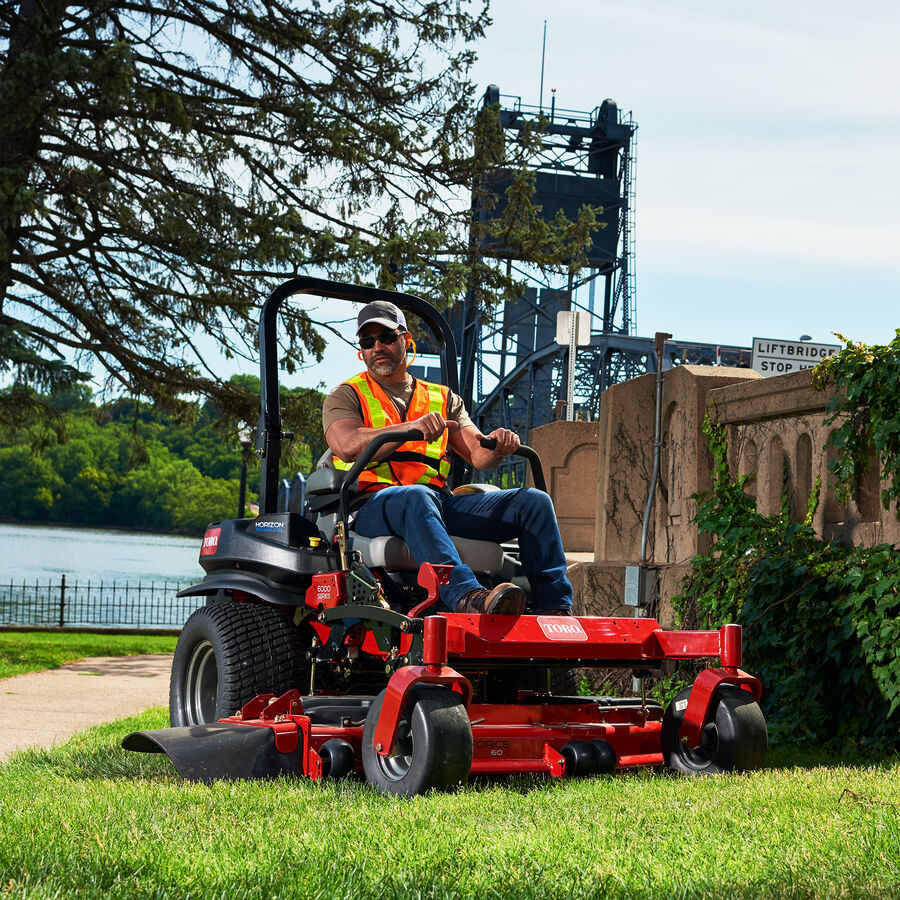 Man on a Toro Z Master 6000 series mower mowing grass. There is a river with a lift bridge in the background.