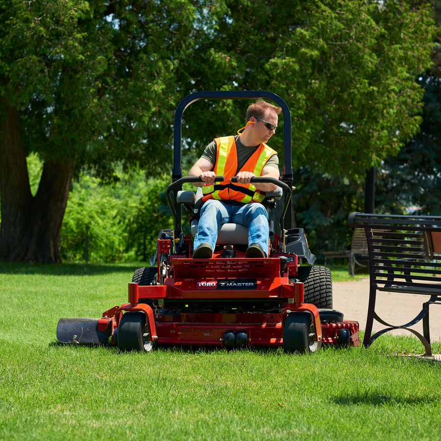 Man on a Toro Z Master 5000 mower mowing grass by a path and a black bench.