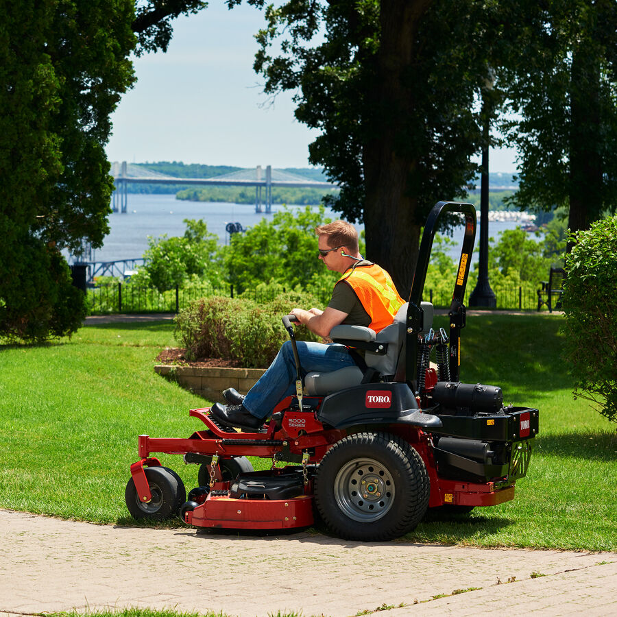 Man mowing grass with a Toro Z Master 5000 mower. They are mowing next to a cement walkway with trees and a river in the background.