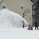 Man using snow blower on driveway in by a basketball hoop
