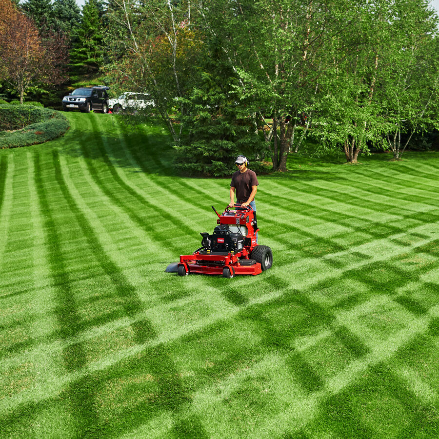 Man using a Toro GrandStand mower mowing an open grass space - trees in the background.  Grass has a checkered pattern.