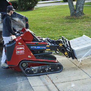 White male operating the Dingo TX 1000 hauling bricks with the forks attachment.