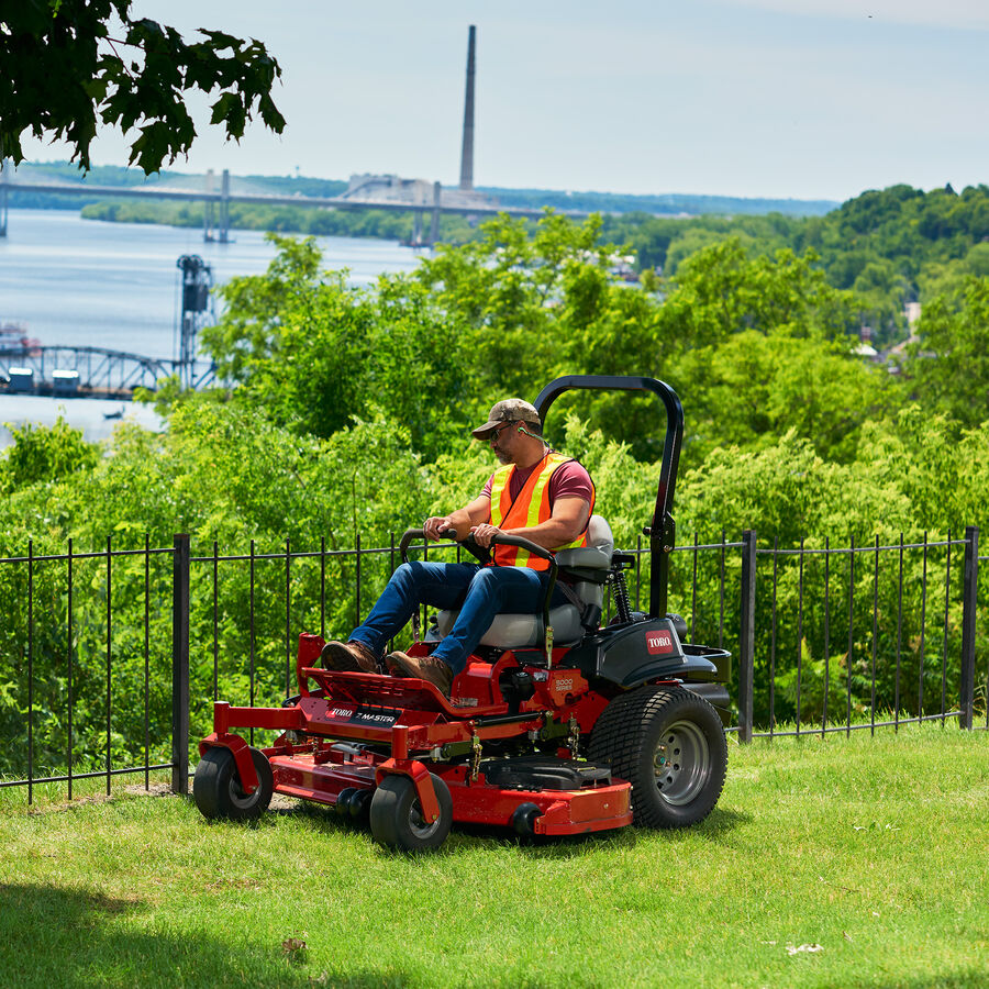 Man on a Z Master 5000 series mower mowing by a black iron fence with trees in background.
