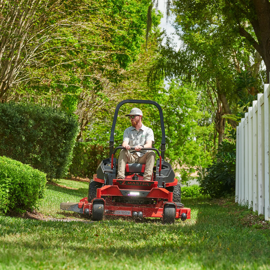 Z Master 2000 Series mower in use - Man on mower mowing lawn between white fence and hedges.