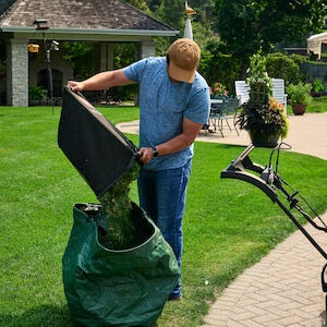 Man dumping grass clippings from the mower's grass catcher bag into a green lawn clippings disposal bag.