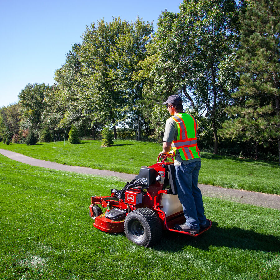 GrandStand® MULTI FORCE Stand-On Gas Mower in use - mowing by a paved walking path.