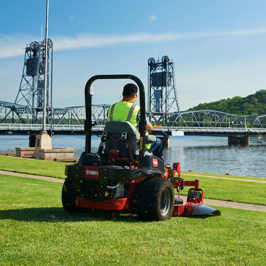 A man on a Toro 6000 Z Master series mower. Mower is on the grass facing a river with a lift bridge.
