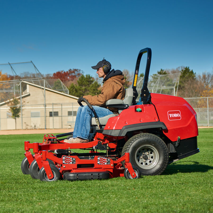 Man on a Toro Z Master 7500 series mower - mowing the outfield of a park baseball field. Baseball field and building are in the background.