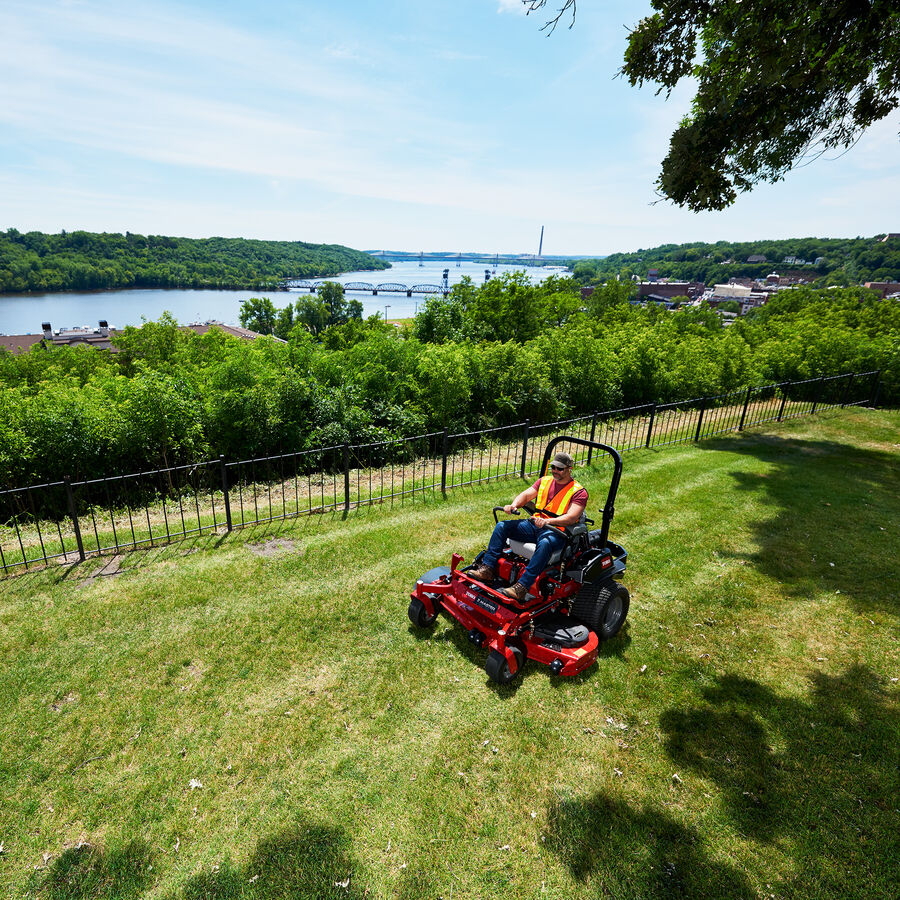 A person in a yellow safety vest mowing with a Toro Z Master 5000 mower on a hillside by a black fence.  A river is in the background.