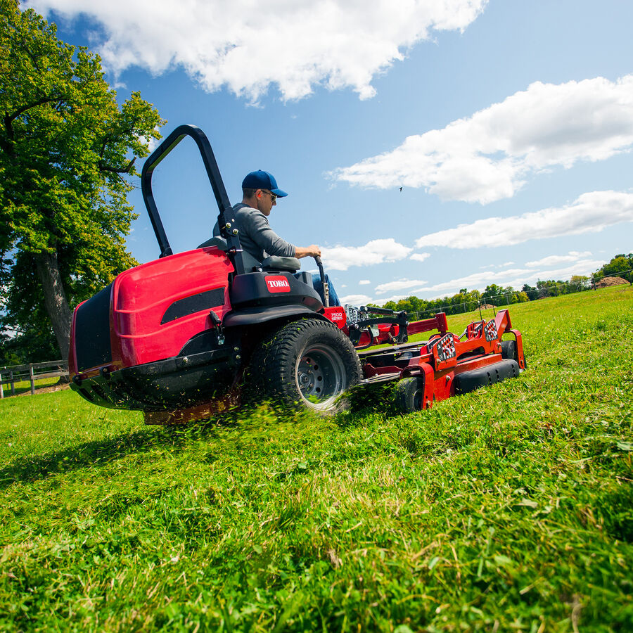 Man on a Toro Z Master 7500 series mower - mowing uphill on a large property with trees in the background.