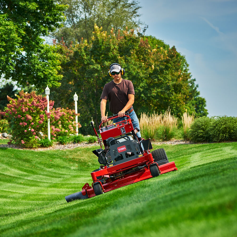 Man using a Toro GrandStand mower mowing on a slight incline with trees in the background
