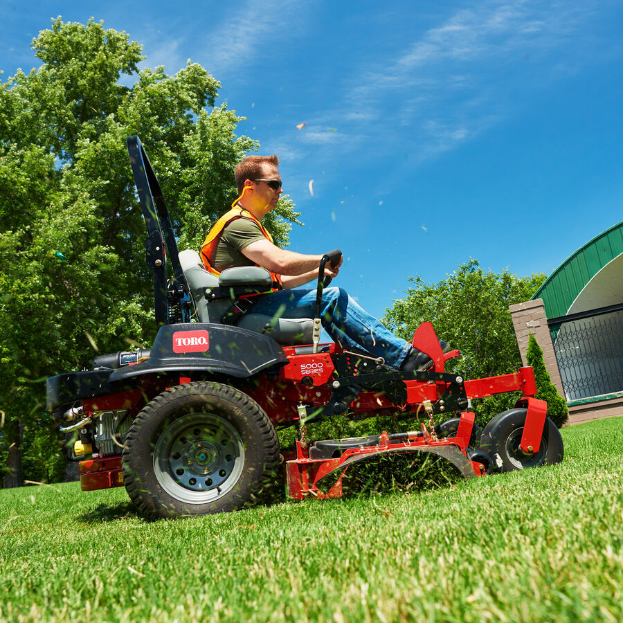 Man on a Z Master 5000 mower mowing grass uphill with a tree in the background
