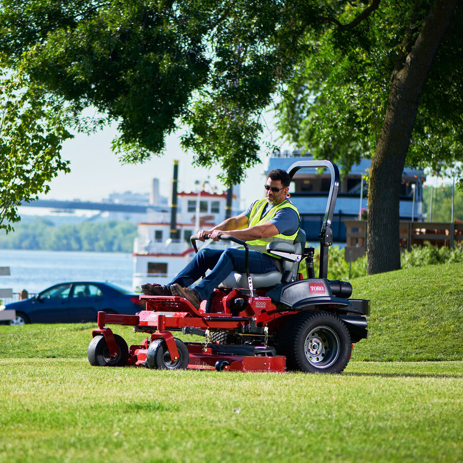 Man mowing grass with a Toro Z Master 6000 mower. Trees, a river and a large boat are in the background. 