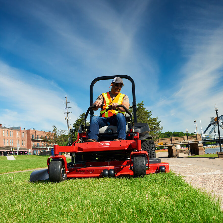 Man mowing with a Toro Z Master 6000 series mower. He is mowing next to a sidewalk, a building to the left and a lift bridge on the right.