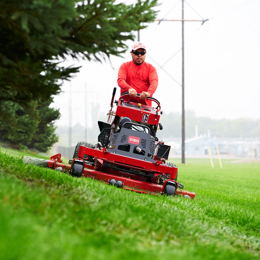 Man using a Toro GrandStand mower mowing on a slight incline with trees to the left