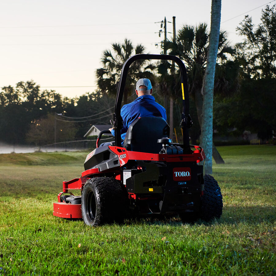 Man on a Z Master 2000 zero turn mower mowing a park lawn with a tree next to the mower and trees in the background.