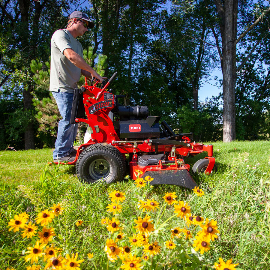 GrandStand® MULTI FORCE Stand-On Gas Mower in use - mowing by some wild yellow daisies